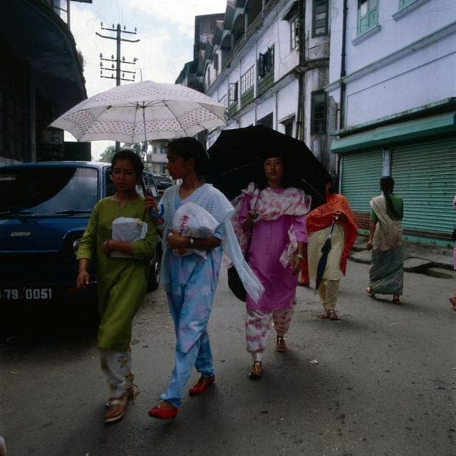ছাতা হাতে রঙিন সালোয়ার-কামিজ পরা কয়েকজন নারী ভারতের দার্জিলিংয়ের একটি রাস্তা দিয়ে হেঁটে যাচ্ছেন। ছবিটি ১৯৮০ সালের দিকে তোলা